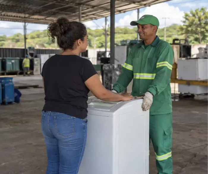 Moradora entrega uma máquina de lavar a um funcionário uniformizado em uma área coberta de recebimento de resíduos.
