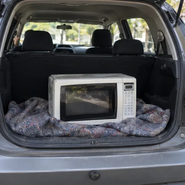 Um micro-ondas usado está apoiado sobre uma manta no porta-malas de um carro para evitar impactos durante o transporte.