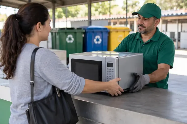 Pessoa entregando um micro-ondas a um atendente em um ponto de coleta com recipientes de reciclagem.