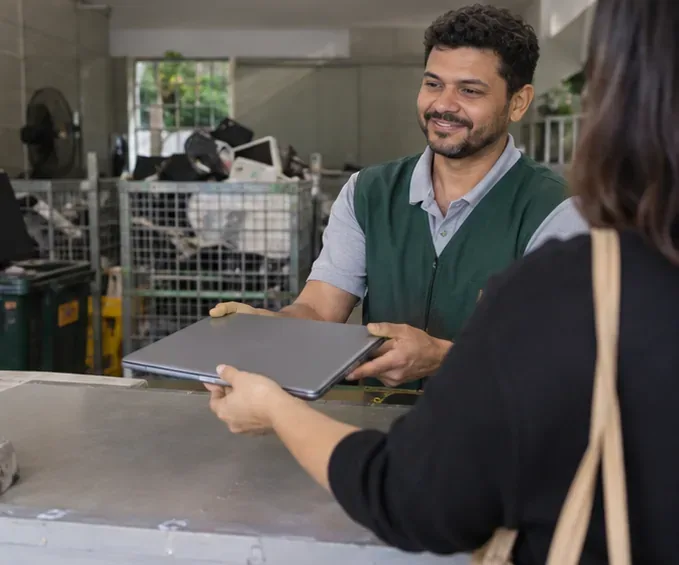 Atendente recebe um notebook fechado de uma moradora em um ponto de coleta de eletrônicos
