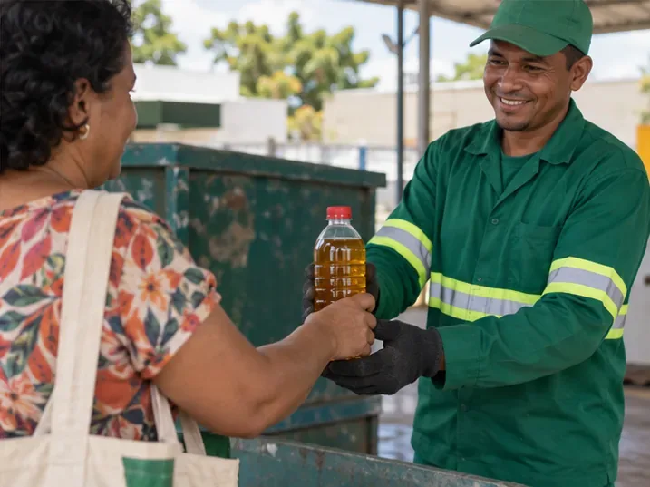 Moradora entrega uma garrafa PET fechada com óleo usado a um funcionário em um ponto de recebimento de recicláveis.