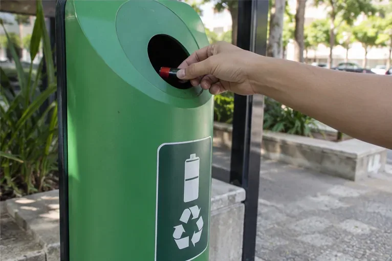 Mão depositando uma pilha em um coletor verde identificado com símbolo de bateria e reciclagem