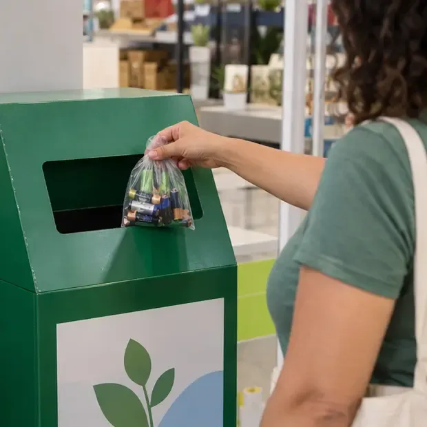 Pessoa entrega um pequeno saco com pilhas usadas em uma caixa de coleta verde dentro de uma área comercial
