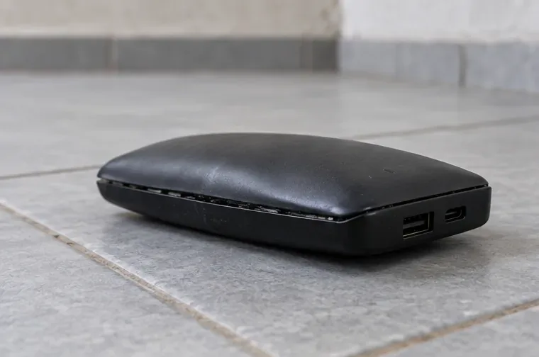 Power bank preto visivelmente estufado e desconectado sobre um piso cerâmico frio, sem ninguém tocá-lo