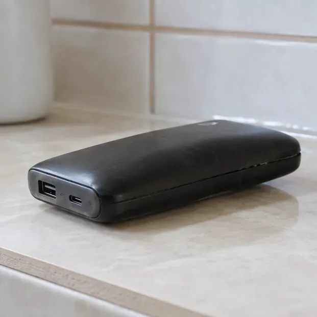 Power bank preto visivelmente estufado apoiado sobre uma superfície de cerâmica, sem cabos conectados