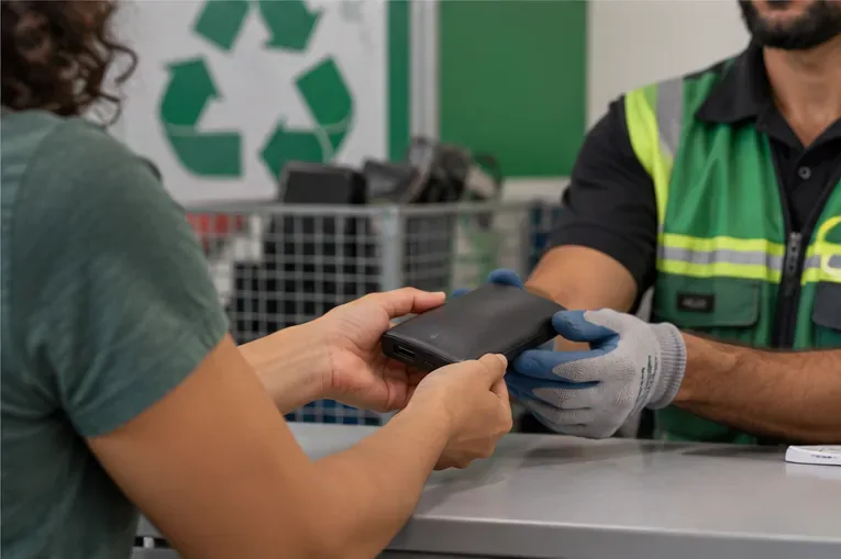 Pessoa entregando um power bank a um funcionário com luvas em um ponto de recebimento de eletrônicos