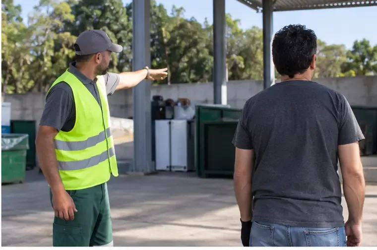 Funcionário usando colete de alta visibilidade indicando a área correta a um morador dentro do Ecoponto