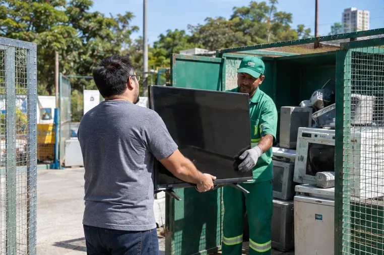 Pessoa entregando uma televisão a um trabalhador em uma área de recebimento de resíduos eletrônicos