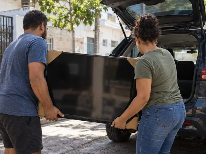 Duas pessoas carregam uma televisão na vertical até um carro, apoiando-a com cuidado
