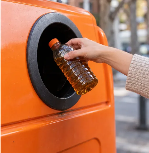 Una mano introduce una botella de plástico cerrada con aceite usado en un contenedor naranja