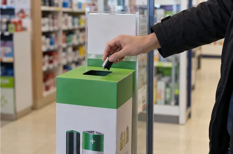Una mano deposita una pila doméstica usada en un pequeño contenedor de recogida situado dentro de un comercio