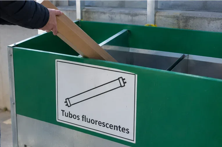 Caja de cartón con un tubo fluorescente siendo introducida cuidadosamente en un contenedor identificado para tubos fluorescentes