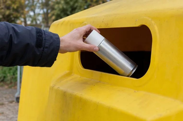 Una mano deposita un aerosol metálico vacío en la abertura de un contenedor amarillo