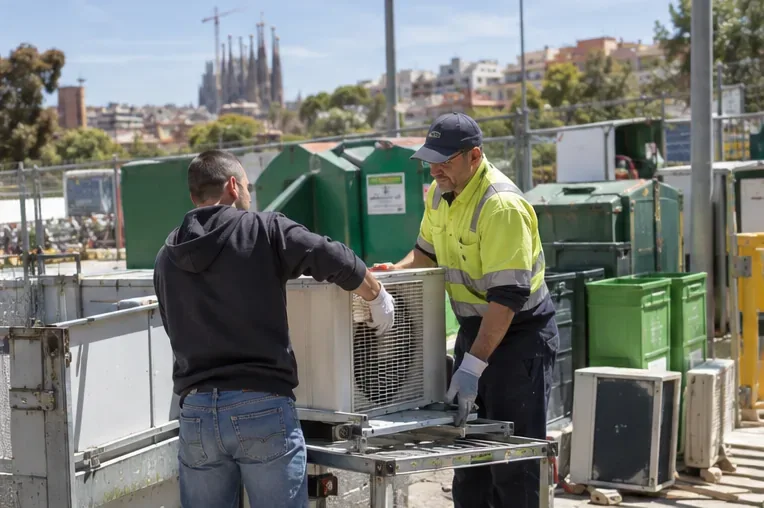 Trabajador de un centro de recogida ayudando a descargar una unidad exterior de aire acondicionado en una zona de recepción de residuos eléctricos