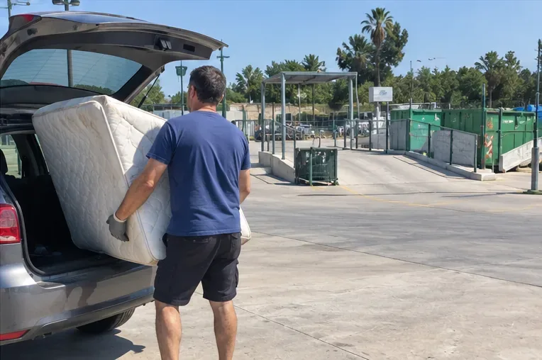 Persona descargando un colchón de un vehículo en una instalación de ecoparque con contenedores de gran tamaño.