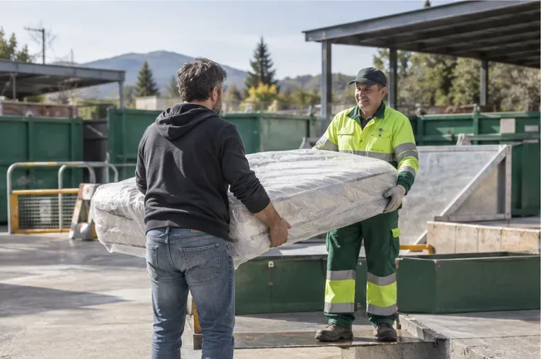 Una persona entrega un colchón protegido a un trabajador en una instalación de reciclaje al aire libre