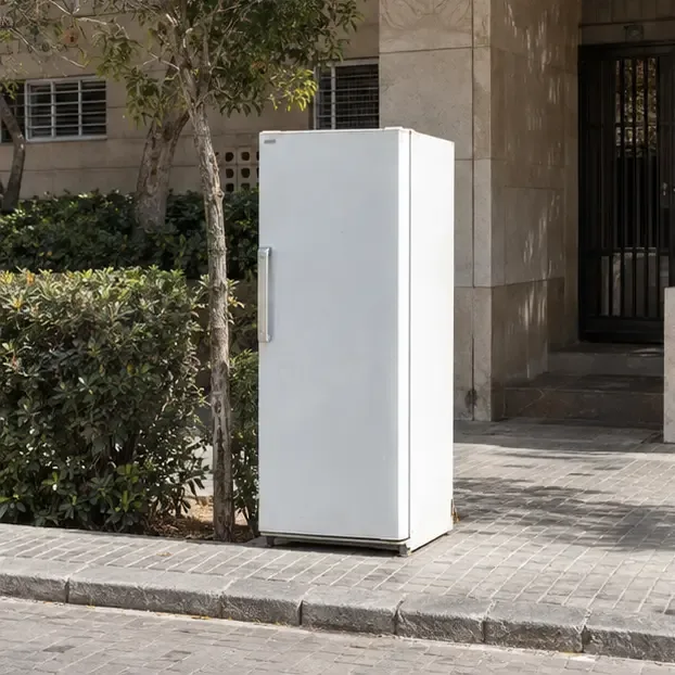 Frigorífico blanco colocado de forma ordenada en un punto de recogida acordado frente a un edificio residencial, sin bloquear la acera.