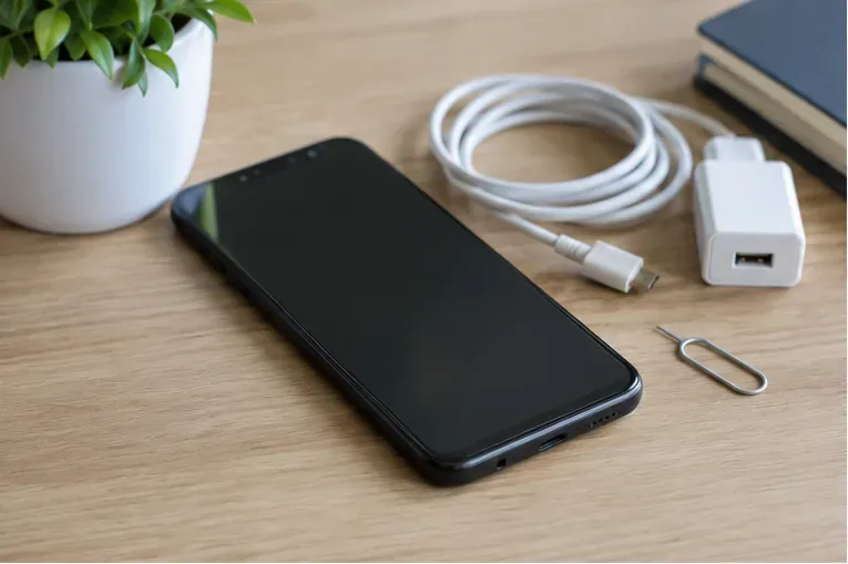 Teléfono móvil sobre una mesa junto a cable de carga y herramienta para extraer la SIM antes de prepararlo para reciclaje