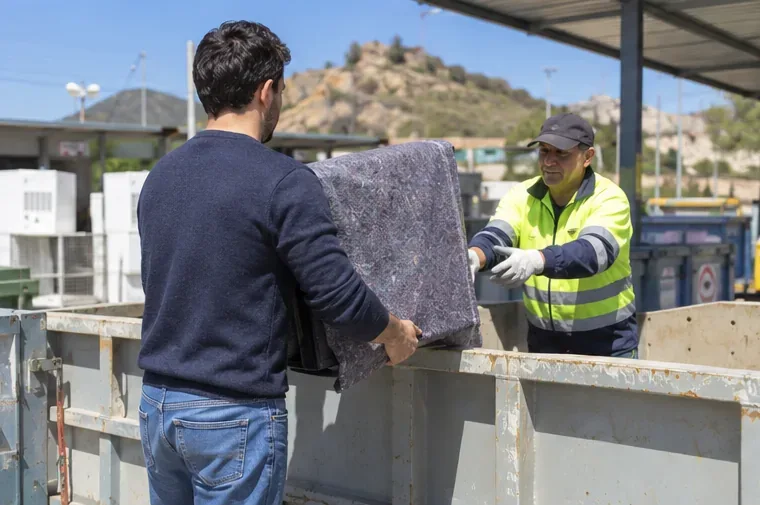 Una persona entrega un televisor protegido a un trabajador de un centro de reciclaje junto a contenedores de residuos