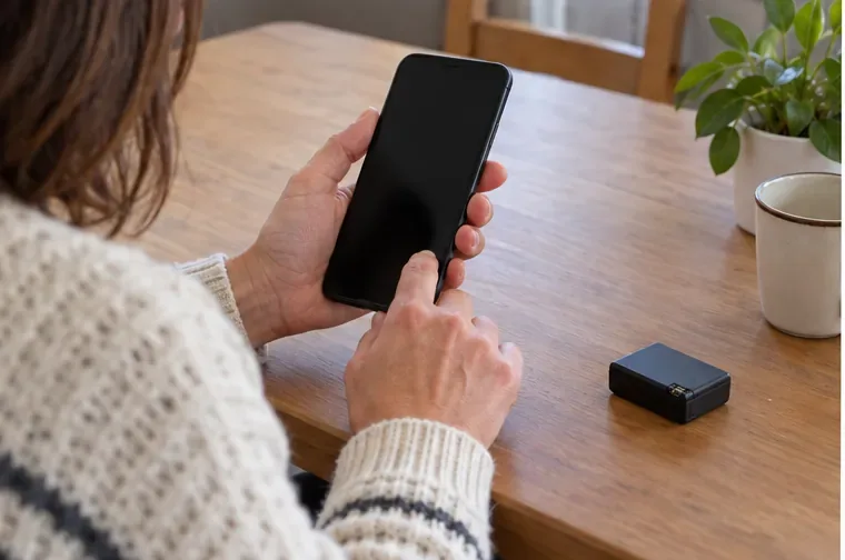 Persona consulta un teléfono con la pantalla apagada junto a una pequeña batería sobre la mesa