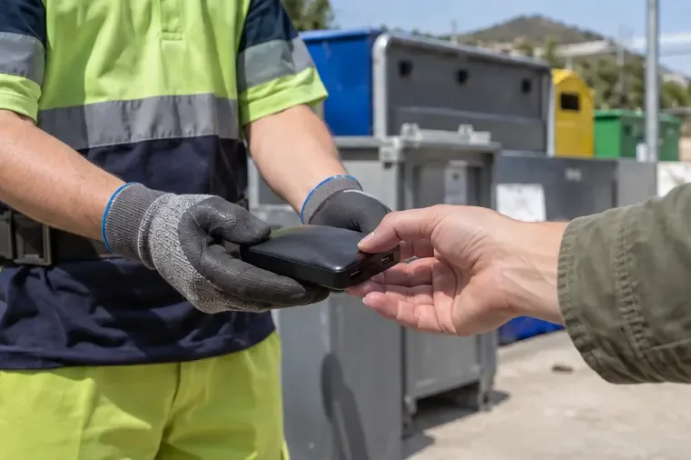 Trabajador con guantes recibe una batería externa de manos de un usuario en una zona de recogida de residuos