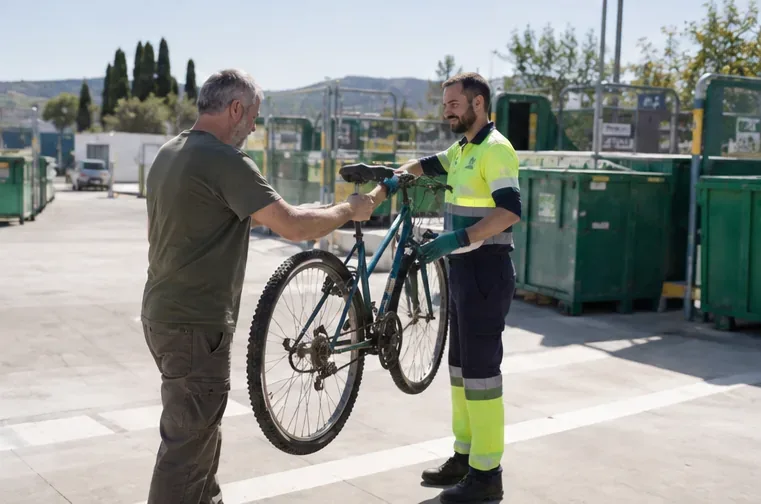 Trabajador con ropa reflectante recibiendo una bicicleta vieja junto a contenedores de residuos separados