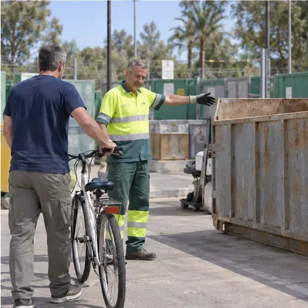 Un trabajador con ropa reflectante indica un contenedor metálico mientras recibe una bicicleta en un área municipal de recogida