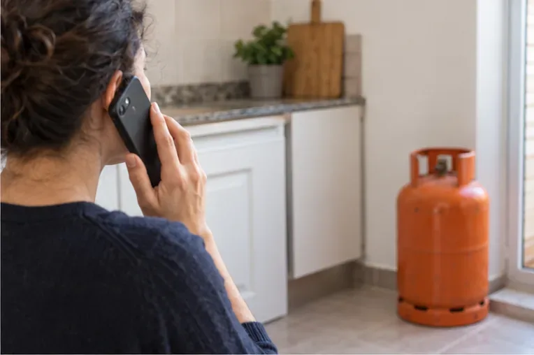 Una mujer llama por teléfono desde una cocina mientras una bombona de gas permanece colocada en vertical a varios metros de distancia.