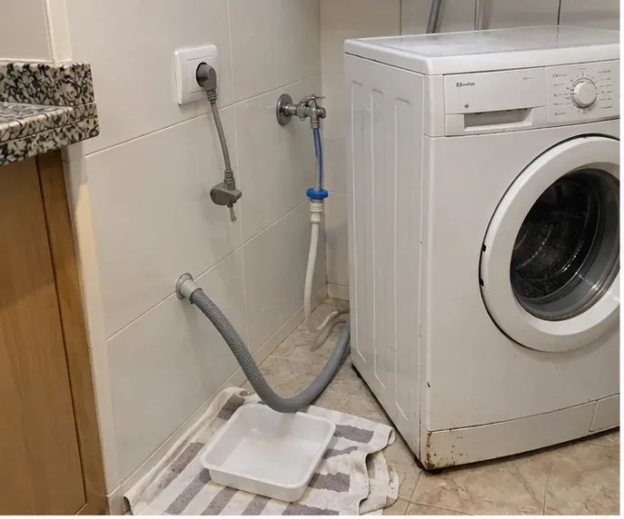 Lavadora desconectada en una zona de lavado, con manguera de desagüe dirigida a una bandeja para vaciar el agua restante