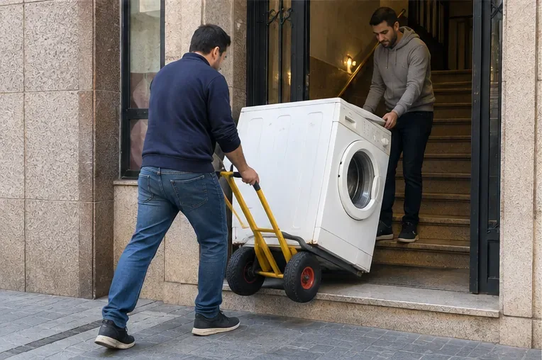 Dos personas bajando una lavadora con una carretilla de transporte por la entrada de un edificio