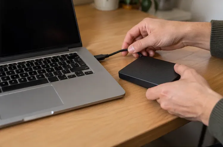 Handen plaatsen een kabel bij een externe schijf naast een open laptop met een donker scherm.