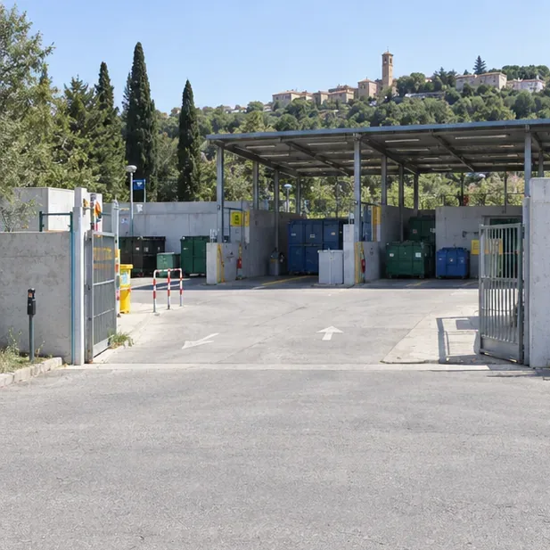 Ingresso di un centro comunale di raccolta con area coperta e contenitori separati, senza insegne leggibili.