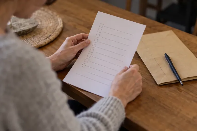 Des mains tiennent une feuille de contrôle au-dessus d’une table, près d’une enveloppe et d’un stylo.