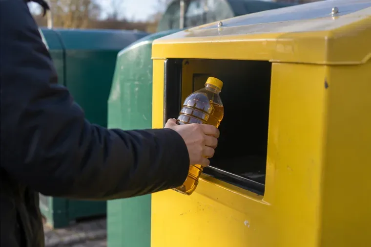 Hand met een afgesloten plastic fles gebruikte frituurolie bij een gele inzamelcontainer