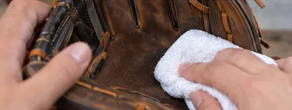 Hands gently wiping the palm of a brown leather baseball glove with a clean white damp cloth
