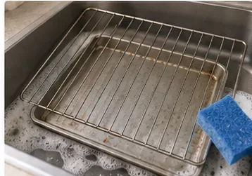 A wire toaster-oven rack and metal pan being washed in warm soapy water with a soft sponge.