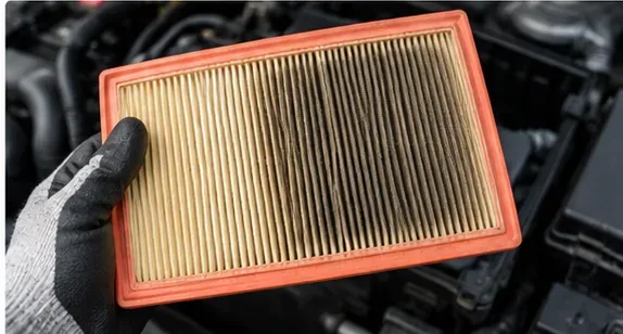 A rectangular engine air filter held above an engine bay, with one side visibly darker from trapped dust