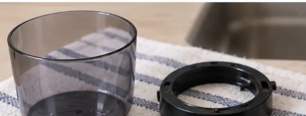 A transparent grinder hopper and black removable plastic ring resting on a clean striped towel beside a sink