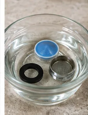 Faucet aerator parts soaking in a clear bowl of cleaning solution on a countertop.