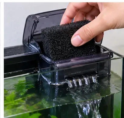 A hand places a cleaned black sponge back into the filter chamber above the aquarium.