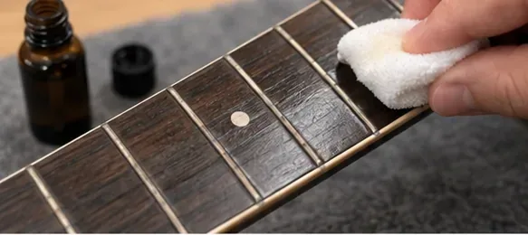 A small cloth spreading a very light coat of fretboard conditioner on an unfinished dark wood guitar fretboard