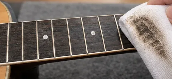A white microfiber cloth wiping dark grime from a guitar fretboard between the metal frets