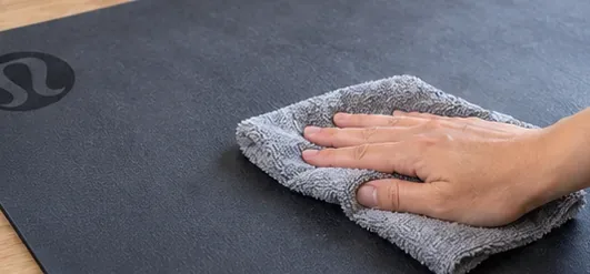 A hand wipes the surface of a dark yoga mat with a soft gray microfiber cloth before applying cleaner.