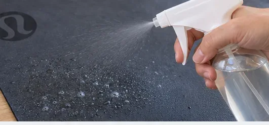 A hand applies a light mist of cleanser to a dark yoga mat without soaking the surface.