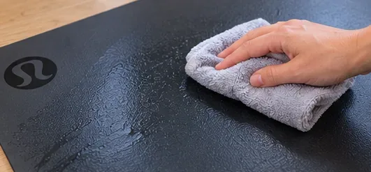 A hand makes a second pass across a dark yoga mat with a clean damp microfiber cloth.