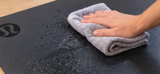 A hand wipes a damp dark yoga mat evenly with a folded microfiber cloth.
