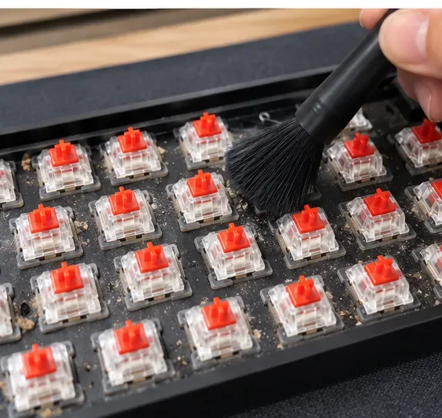 A soft black detailing brush sweeping crumbs and dust from the plate of a mechanical keyboard with clear switch housings and red stems.