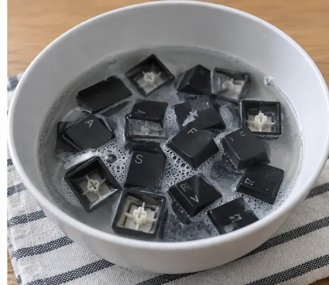 Black mechanical keyboard keycaps soaking in a white bowl of warm soapy water with small bubbles around the caps.