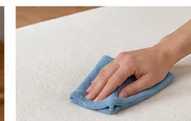 A hand blotting a small stain on a white mattress topper with a blue microfiber cloth