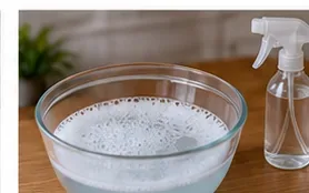 A glass bowl containing a small amount of sudsy cleaning solution beside a microfiber cloth and spray bottle
